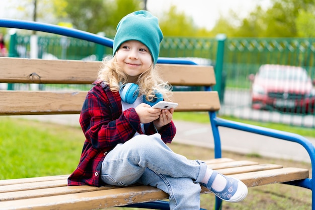 Garotinha loira com smartphone nas mãos e fones de ouvido, sentada em um banco no parque.