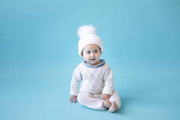 Garotinha loira com um chapéu de malha branco de inverno e uma camisola de malha em um fundo azul isolado, espaço para texto, criança pequena em roupas de inverno