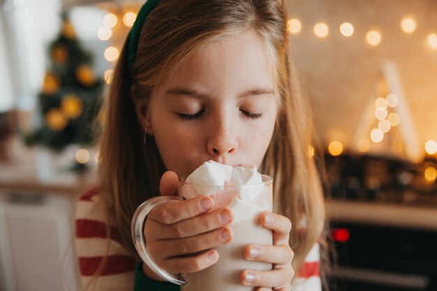 Garotinha loira de pijama listrado segura uma caneca com bebida quente e marshmallows na cozinha decorada. fins de semana e feriados de inverno. lugar para texto.