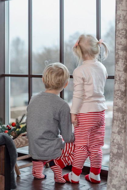 Garotinha loira e menino ficam perto da janela Irmão mais velho com irmã Vista traseira Quadro vertical da véspera de Natal