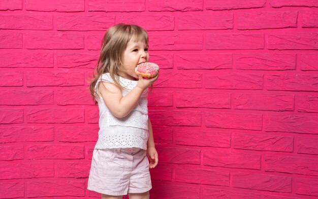 Garotinha loira engraçada em camisa branca come donut na parede rosa.