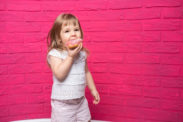 Garotinha loira engraçada em camisa branca come donut no fundo rosa.