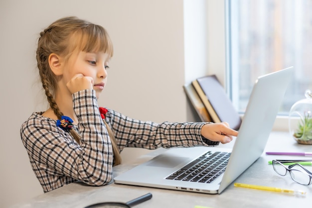 Garotinha loira fazendo lição de casa em casa à mesa. A criança é educada em casa. Uma garota de cabelo claro executa uma tarefa online usando um laptop e um tablet.