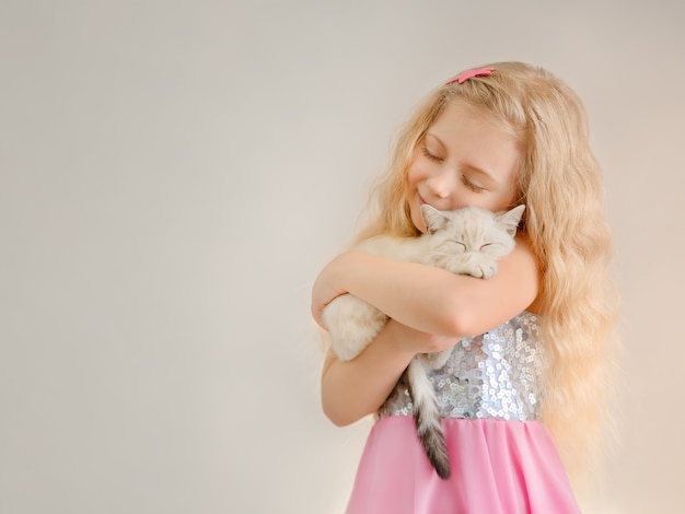 Garotinha loira feliz abraça um gatinho escocês bege adormecido e sorri fechando os olhos em um vestido brilhante