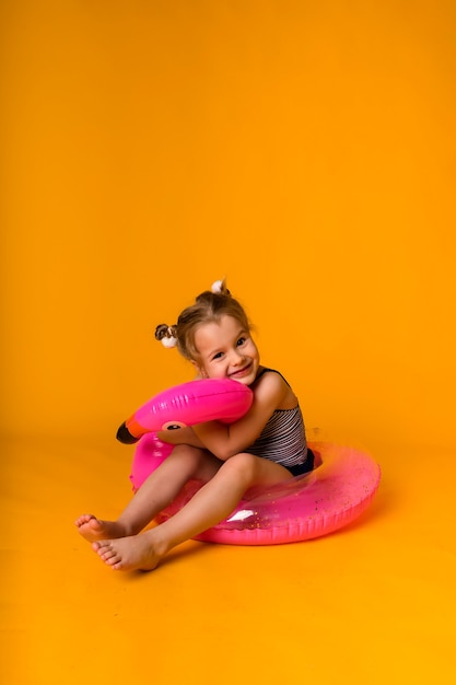 Garotinha loira feliz em um maiô listrado senta-se em um círculo inflável rosa e abraça um flamingo em um fundo amarelo com uma cópia do espaço