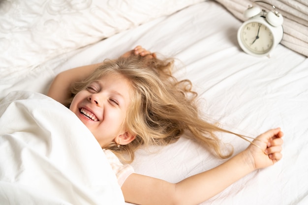 Garotinha loira feliz em uma cama branca com um despertador bom dia