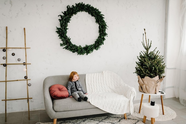 Garotinha sentada em um sofá perto da árvore de Natal em casa Os feriados do ano novo de 2021 e o conceito de infância Feliz Natal e Boas Festas
