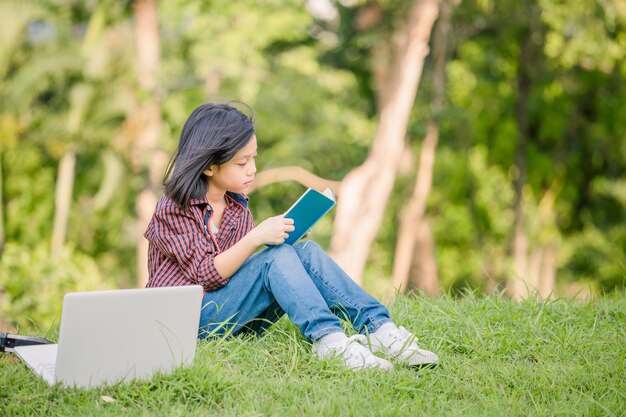 Garotinha sentada na grama e lendo o livro no parque Crianças brincando de conceitos de aprendizagem ao ar livre