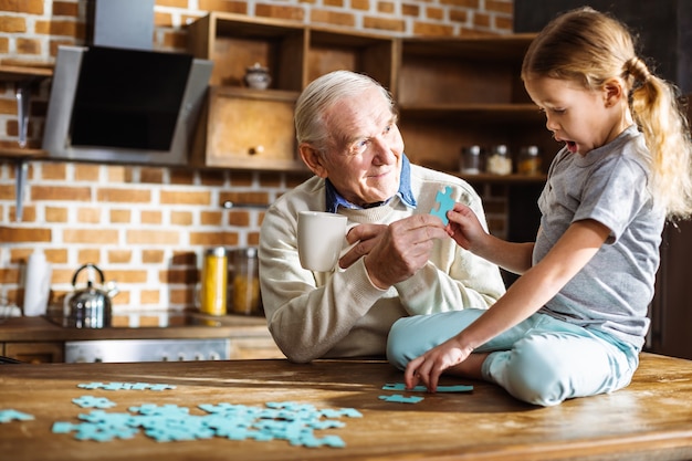 Garotinha simpática sentada na mesa enquanto monta o quebra-cabeça com o avô