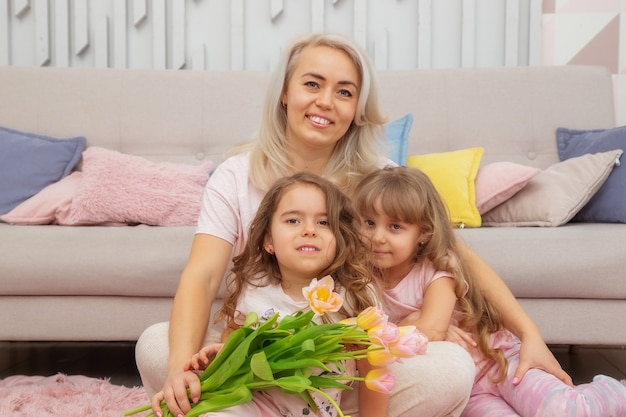 Garotinhas de aparência caucasiana se abraçam à mãe, sentadas no chão em frente ao sofá em uma sala bem iluminada em estilo escandinavo