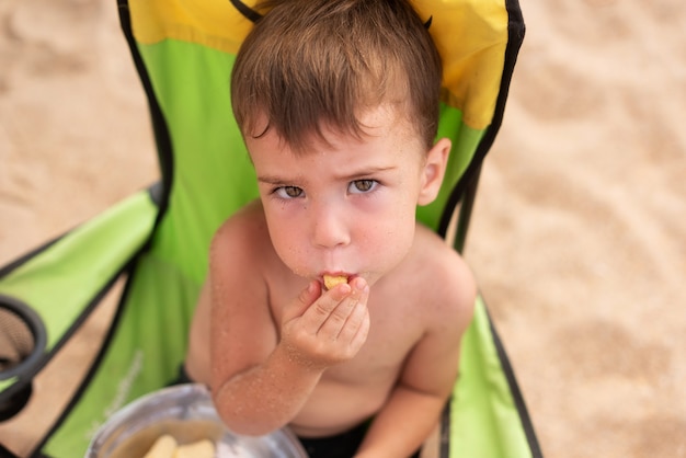 Garotinho adorável comendo palitos de milho em uma tigela de lata na praia perto do mar