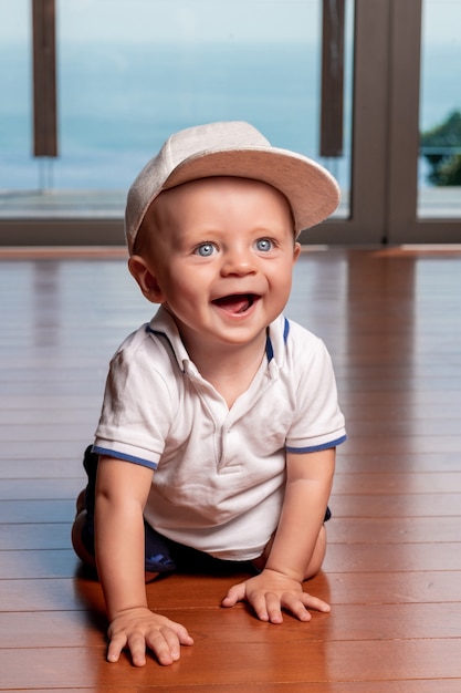 Garotinho com boné cinza e descalço, rastejando no chão, sorrindo e olhando para o lado