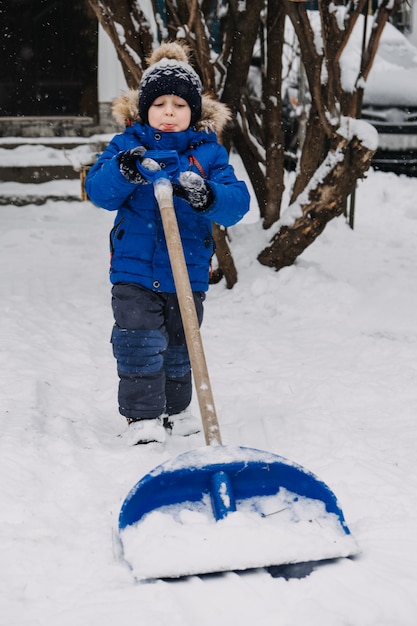 Garotinho com uma pá na mão remove a neve no quintal garoto de jaqueta azul limpa a neve