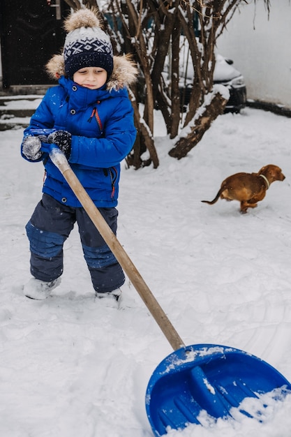 Garotinho com uma pá na mão remove a neve no quintal Garoto de jaqueta azul limpa a neve