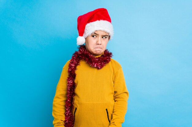 Garotinho comemorando o dia de Natal usando um chapéu de Papai Noel isolado confuso, sente-se duvidoso e inseguro.