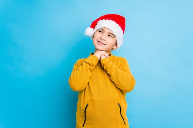 Garotinho, comemorando o dia de Natal, usando um chapéu de Papai Noel isolado mantém as mãos sob o queixo, está olhando alegremente de lado.