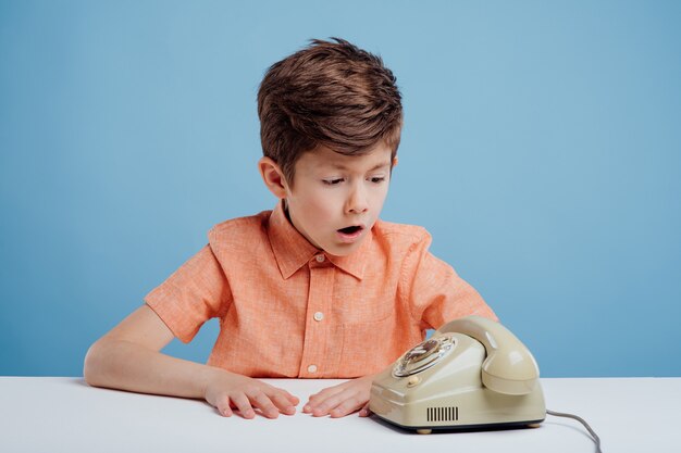 Garotinho confuso com telefone antigo sentado à mesa fundo azul