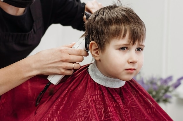 Garotinho cortado com máquina de cabeleireiro Closeup de mãos de mulher preparando o cabelo de garoto na barbearia Início do corte de cabelo Retrato de criança do sexo masculino na barbearia para cortar o cabelo