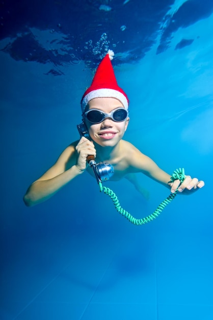 Garotinho de boné vermelho de Papai Noel nada e posa debaixo d'água na piscina com telefone na mão