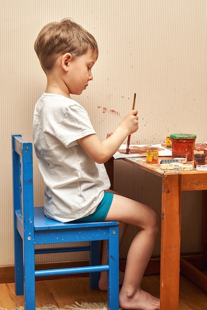 Garotinho desenha com pincel e cor marrom na mesa com potes sujos e brinquedos