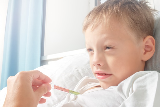 Garotinho doente na cama com a temperatura enquanto sua mãe está medindo sua temperatura. O garoto pegou um resfriado. Cuidados de saúde, gripe, higiene.