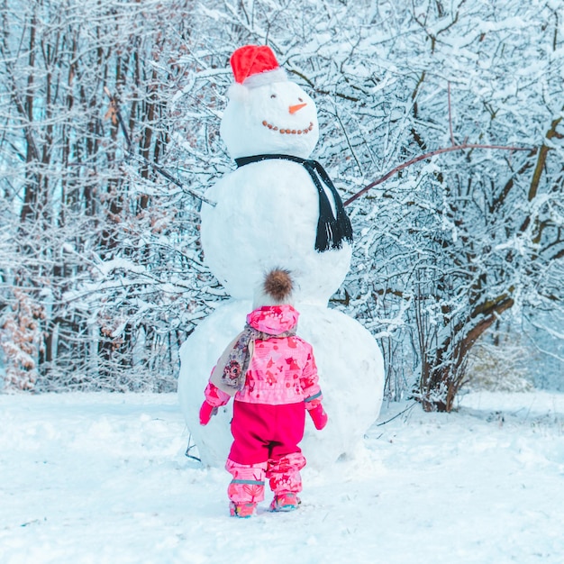 Garotinho dourado com roupas de inverno rosa, olhando para o grande boneco de neve. inverno