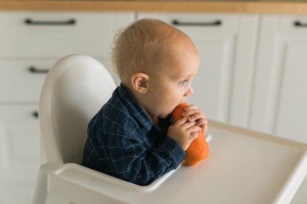Garotinho em uma camiseta azul sentado em uma cadeira de criança comendo cuidados com o bebê de cenoura e alimentação infantil
