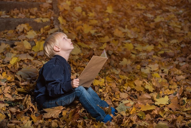 Garotinho está sentado na floresta selvagem em folhas de outono com livro nas mãos Livros infantis sobre milagres e magia