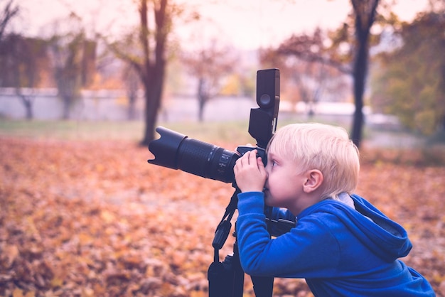 Garotinho loiro atira com uma grande câmera SLR em um tripé Sessão de fotos no parque de outono