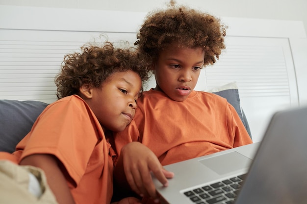 Garotinho olhando como seu irmão joga no laptop quando estão sentados na cama