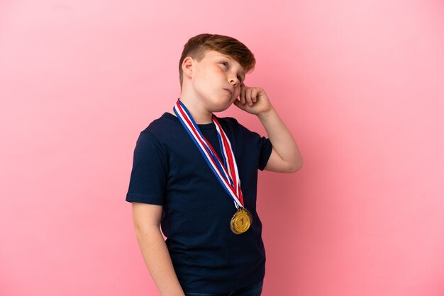 Garotinho ruivo com medalhas isoladas em fundo rosa tendo dúvidas e com expressão facial confusa