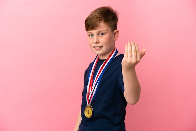 Garotinho ruivo com medalhas isoladas em um fundo rosa, convidando para vir com a mão. Feliz que você veio