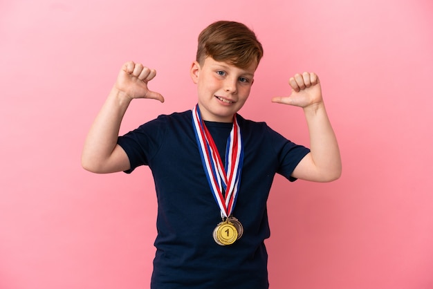 Garotinho ruivo com medalhas isoladas em um fundo rosa orgulhoso e satisfeito