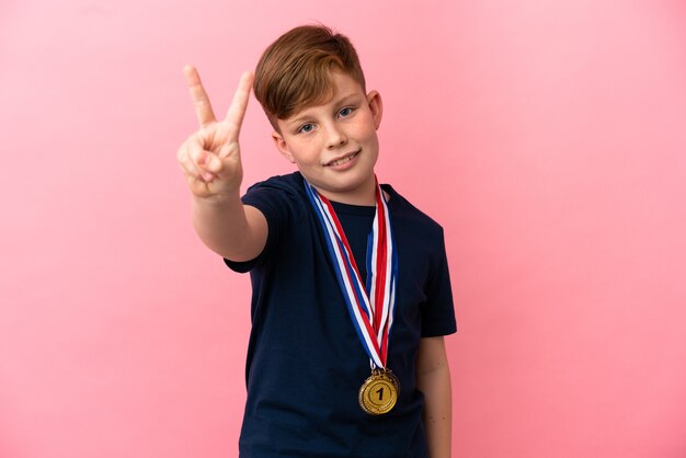 Garotinho ruivo com medalhas isoladas em um fundo rosa sorrindo e mostrando sinal de vitória