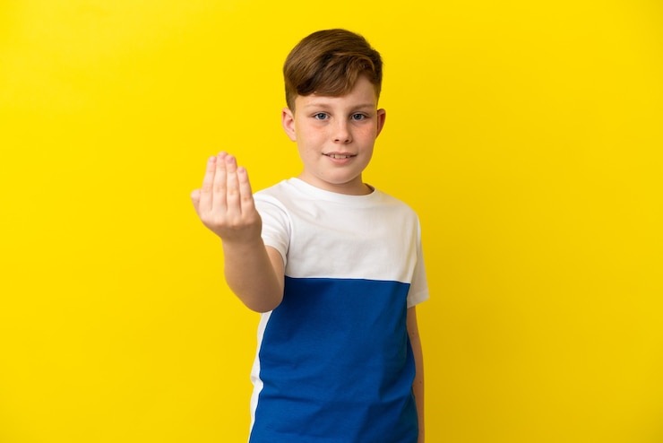Foto garotinho ruivo isolado em um fundo amarelo, convidando para vir com a mão. feliz que você veio