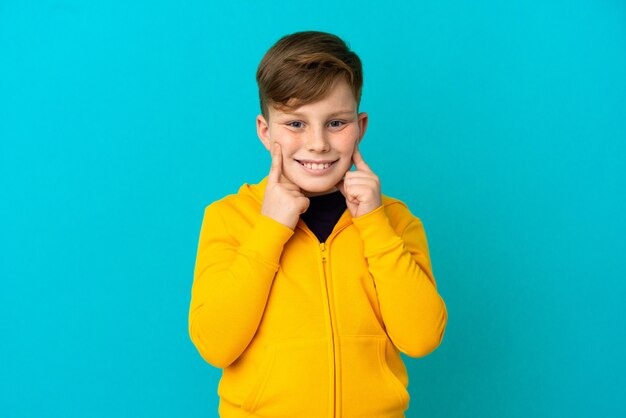 Garotinho ruivo isolado em um fundo azul sorrindo com uma expressão feliz e agradável