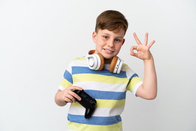 Garotinho ruivo segurando um gamepad isolado na parede branca e mostrando o sinal de ok com os dedos