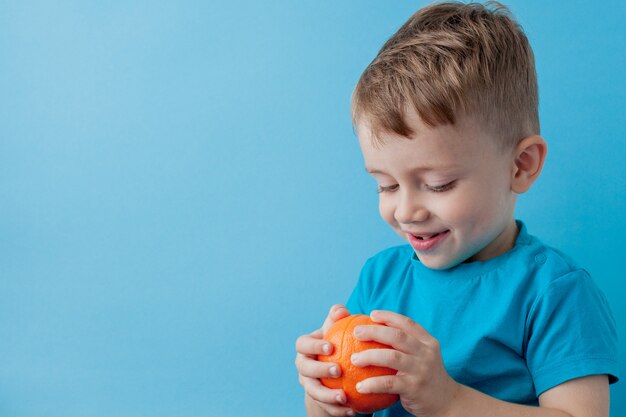 Garotinho segurando uma laranja nas mãos sobre fundo azul, dieta e exercícios para o conceito de boa saúde
