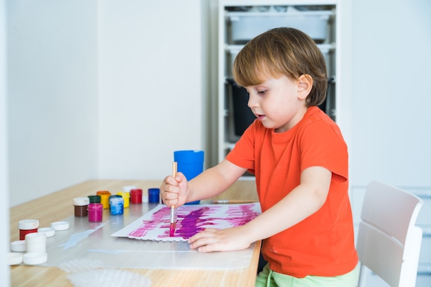 Garotinho sentado à mesa fazendo desenhos coloridos com pincéis