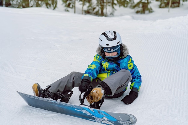 Garotinho sentado na neve colocando os pés em fixações de snowboard ajustando as tiras