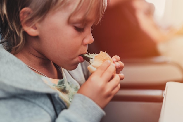 Garotinho sincero menino de cinco anos come hambúrguer ou sanduíche sentado no assento do avião em voo viajando do aeroporto crianças dão uma mordida no avião comendo almoço ou jantar refeição flare