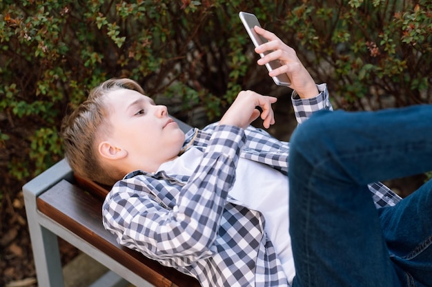 Garoto adolescente atraente e pensativo descansando deitado no banco segurando e usando smartphone para networking