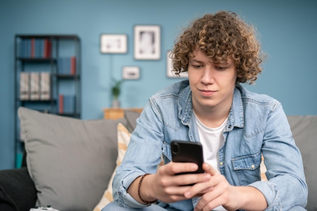 Garoto adolescente atraente sorridente sentado no sofá em casa conversando com um amigo pelo smartphone usando o vídeo c