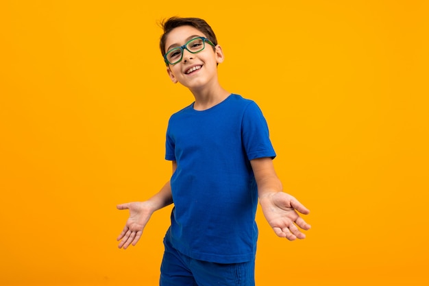 Garoto adolescente bonito em uma camiseta azul com óculos espalha as mãos sobre um fundo amarelo com espaço de cópia