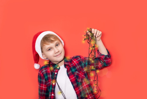 Garoto adolescente caucasiano bonito com chapéu de papai noel segura guirlanda de natal em fundo vermelho copiar espaço para design ou texto banner modelo de maquete de ano novo de natal