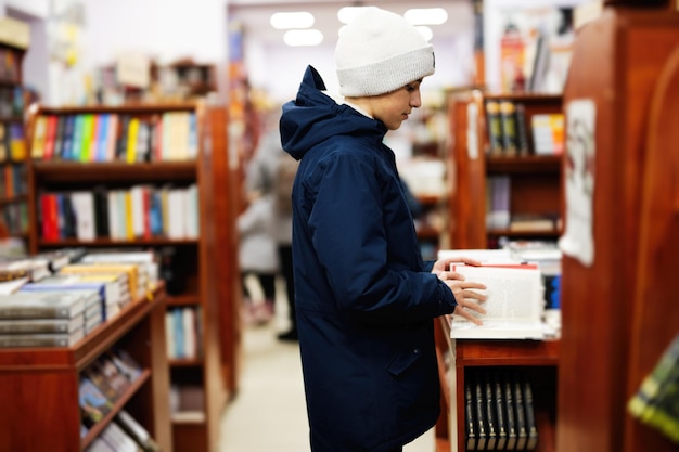 Garoto adolescente de jaqueta alcançando um livro da estante na biblioteca Aprendizagem e educação de crianças europeias