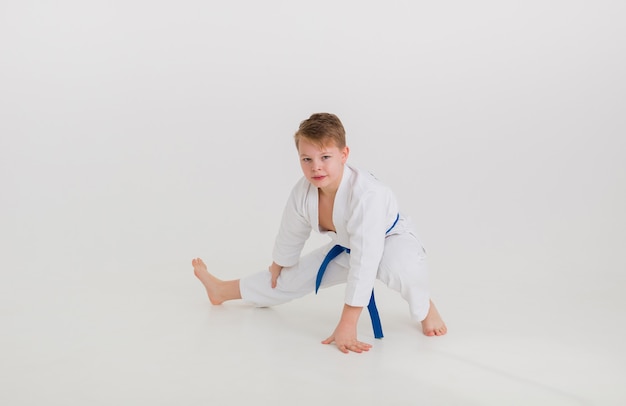 Garoto adolescente em um quimono branco com uma faixa azul se alongando em uma parede branca