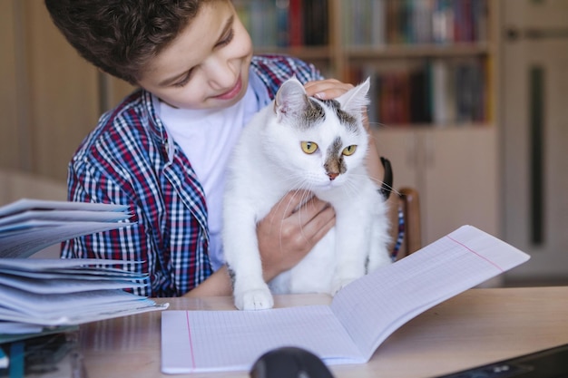 Garoto adolescente em uma mesa segura um gato branco em suas mãos Coronavirus em quarentena escolar em casa Foco seletivo Fundo desfocado