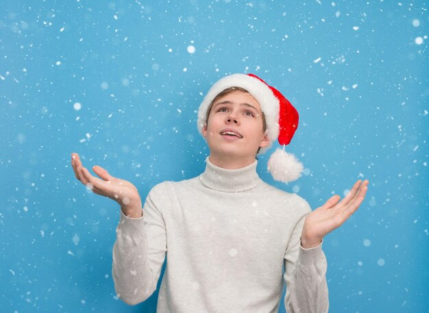 Garoto adolescente feliz com chapéu de Papai Noel pegando flocos de neve no fundo azul nevado