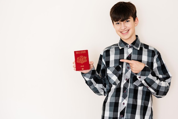 Garoto adolescente segurando o passaporte da Letônia parecendo positivo e feliz de pé e sorrindo com um sorriso confiante contra o fundo branco